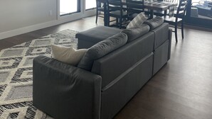 Un amplio salón con un sofá seccional gris, una mesa de comedor con sillas y ventanas grandes que ofrecen vistas al exterior.