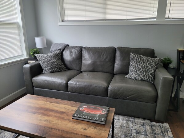 A grey leather sofa with patterned cushions, a wooden coffee table with a magazine, and a grey rug. - Nice and cozy 1 bedroom apartment unit (Ypsilanti)