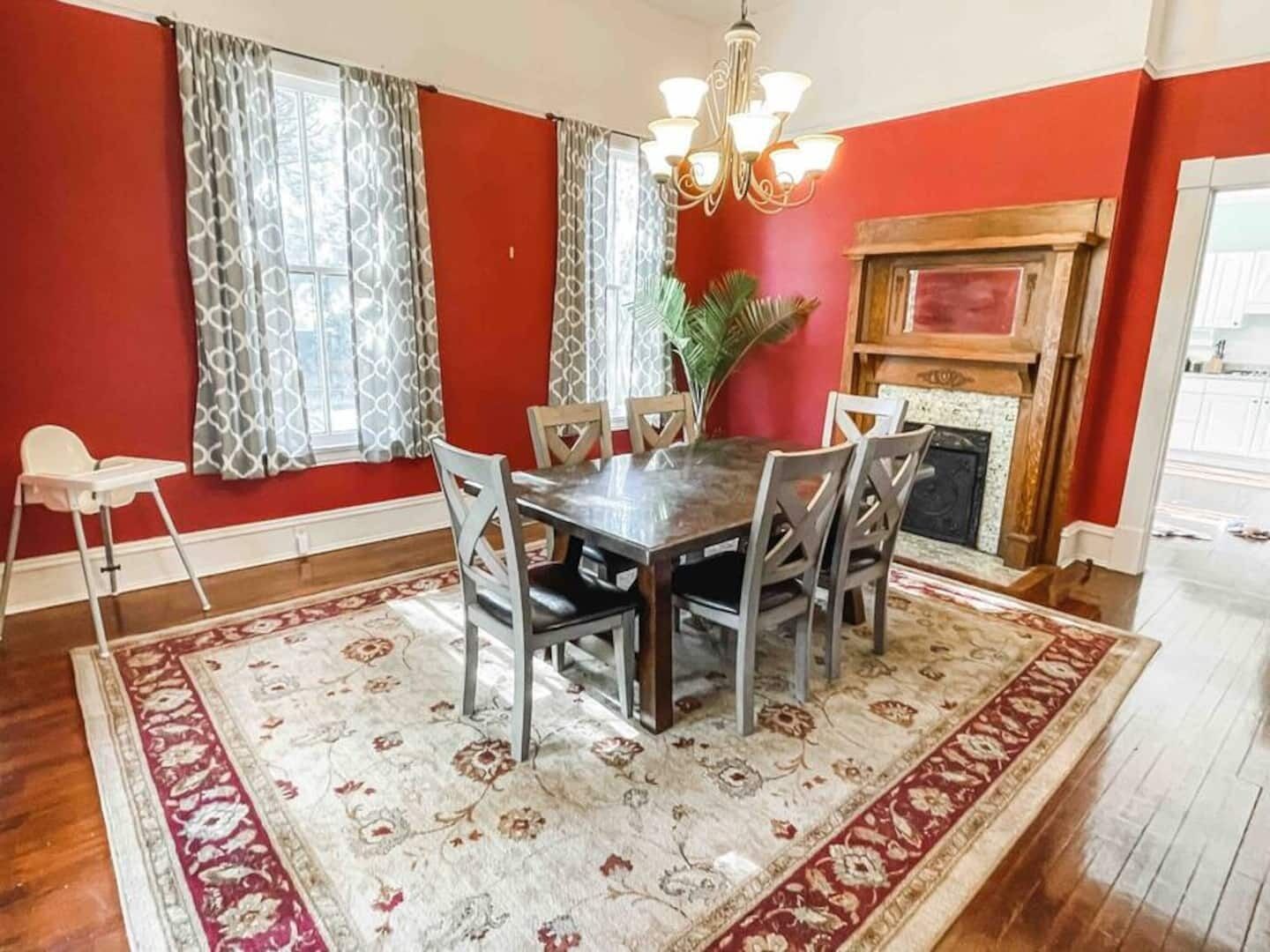 A dining room with a wooden table, six chairs, a chandelier, a fireplace, and a large floral rug.