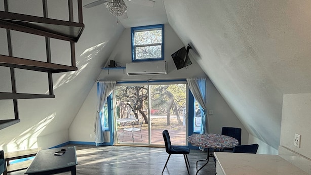 A spacious attic room with a large window, a dining table, chairs, and a kitchenette.