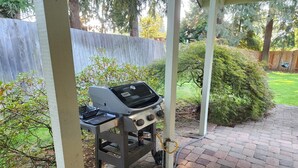 A barbecue grill under a covered patio area with a brick floor and a wooden fence in the background. - Forest Home in quiet cul-de-sac (Eugene)