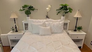 A neatly made bed with a white duvet, two bedside tables with lamps, and two potted plants on either side. - Forest Home in quiet cul-de-sac (Eugene)