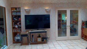 A living room with a flat-screen TV, a wooden entertainment unit, a glass door leading to a balcony, and a tiled floor. - Forest Home in quiet cul-de-sac (Eugene)