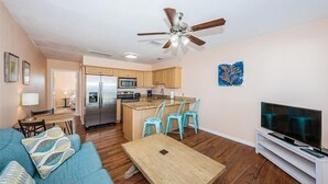 A modern living room with a sofa, a coffee table, and a TV. There is a kitchen area with a refrigerator, microwave, and island with bar stools. A ceiling fan is visible.