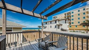 A wooden deck with two Adirondack chairs, overlooking a beach and a multi-story building with balconies.