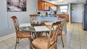 Een eetgedeelte met een glazen tafel, omringd door houten stoelen, een plafondventilator en een keuken op de achtergrond.