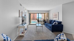 En opholdsstue med sofa, sofabord, fjernsyn og en balkon med udsigt til poolen.