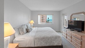 Une chambre à coucher comprenant un lit, des lampes de chevet, une commode, un téléviseur et une fenêtre donnant sur des immeubles et une plage.