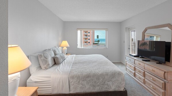 A bedroom with a bed, bedside lamps, a dresser, a television, and a window with a view of buildings and a beach.