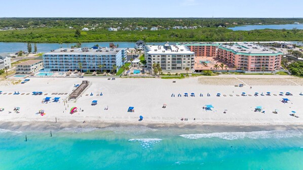 A beachfront hotel with multiple buildings, a sandy beach with umbrellas, and a clear view of the ocean.