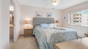 A bedroom with a bed, two bedside lamps, a ceiling fan, and a window with a view of buildings.