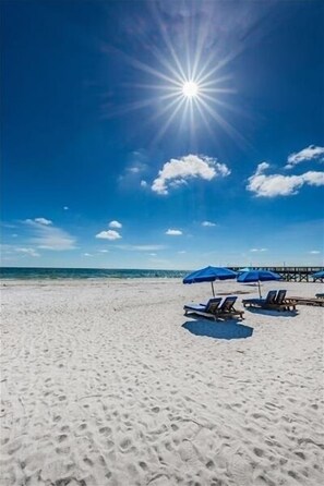 En strand med hvidt sand, klarblå himmel og en strålende sol. Blå parasoller og liggestole er placeret på stranden.