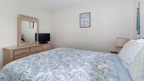Ein Schlafzimmer mit einem Bett, einem Schminktisch, einem Fernseher, einem gerahmten Bild an der Wand und einer Lampe.