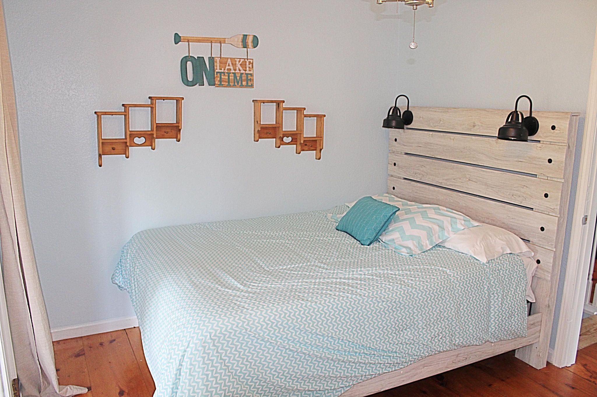 Une chambre avec un lit en bois, une couvre-lit bleue à motifs et une étagère en bois fixée au mur, sur laquelle est posé un panneau indiquant « EN MODE RELAXATION AU LAC ».