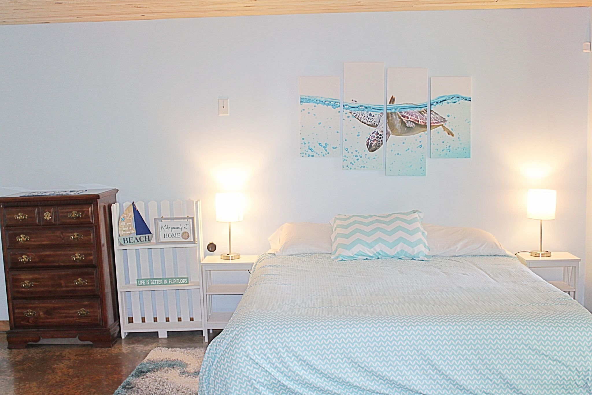 Une chambre à coucher comprenant un lit, des tables de chevet, une commode et un mur orné d’un tableau représentant un paysage marin.