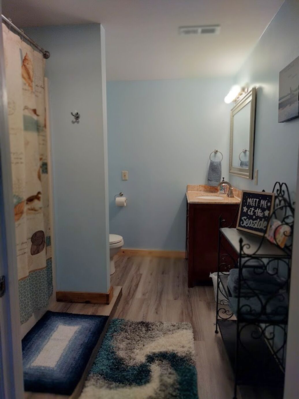 A bathroom with a toilet, sink, and shower area. There is a sign on the wall that reads "MEET ME at the Seaside."