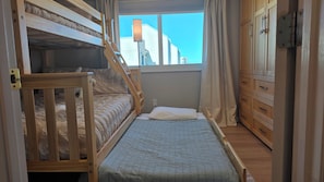 A room with a bunk bed, a single bed, a window with curtains, and wooden cabinets.