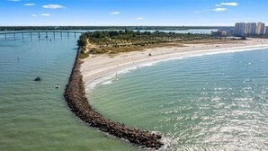 Zona costera con una larga y estrecha península que se adentra en el agua, una playa de arena y un puente al fondo.
