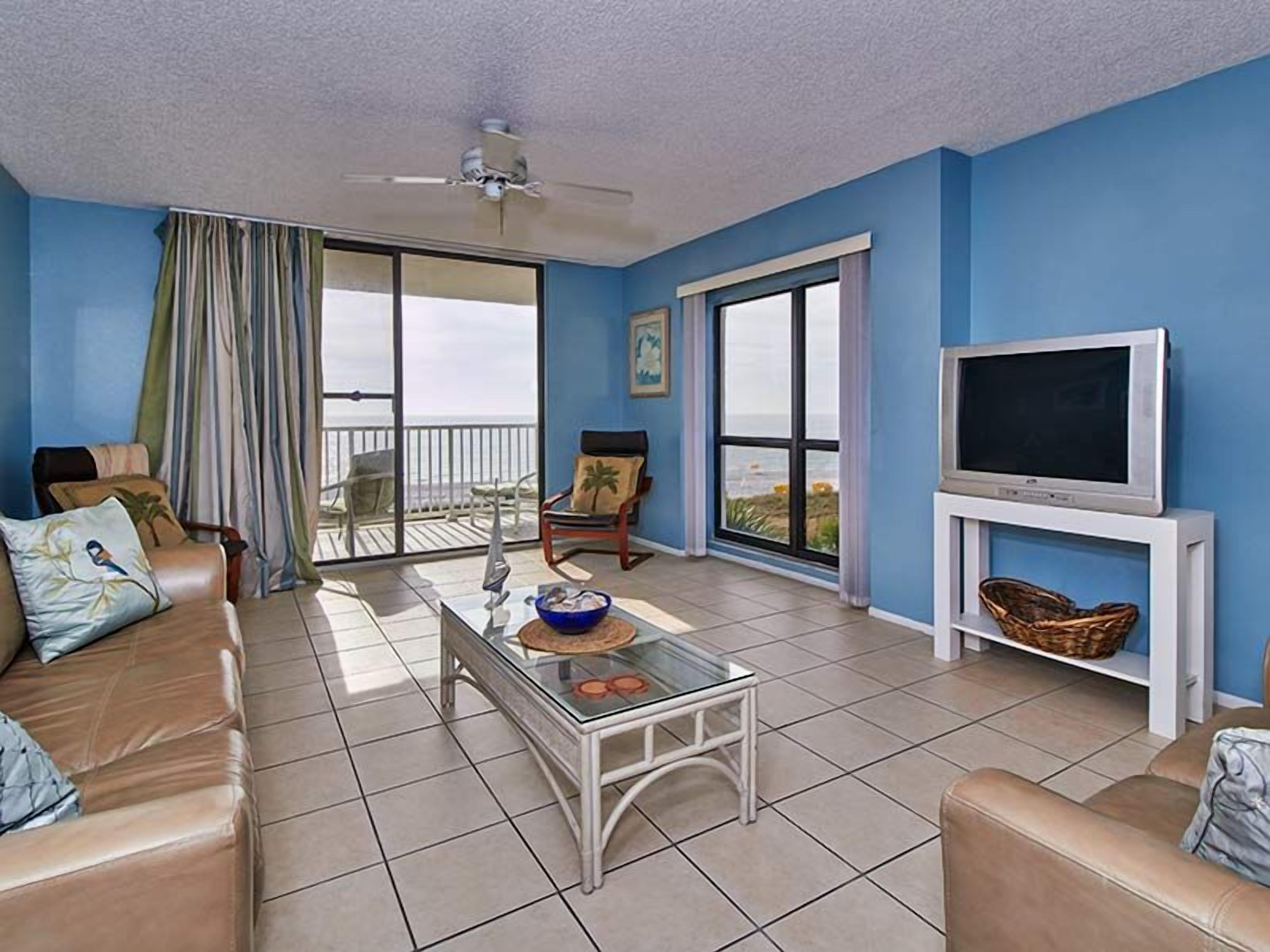 A living room with a sofa, a coffee table, a TV on a stand, and a balcony with a view.