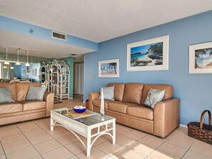 A living room with two leather sofas, a glass coffee table, and framed beach paintings on the wall.