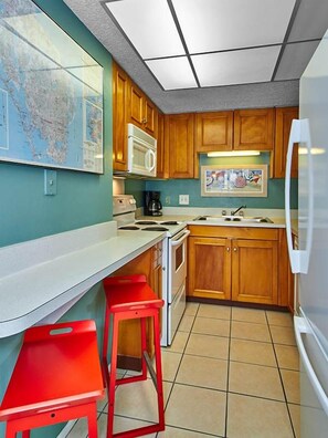 A compact kitchen with wooden cabinets, a white stove, a microwave, and a refrigerator. There are two red bar stools and a map on the wall.