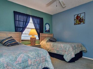 A bedroom with two beds, a window with curtains, a lamp, a mirror, and a painting on the wall.