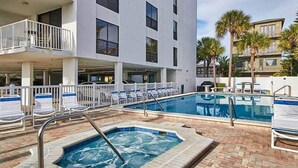 Zona de piscina de hotel con jacuzzi, sillas de descanso y un edificio con balcones.