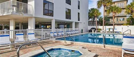 Zona de piscina de hotel con jacuzzi, sillas de descanso y un edificio con balcones.