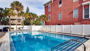 A swimming pool with a metal ladder, surrounded by a white fence, palm trees, and a red building in the background. - Gulf Front Corner Unit & Million Dollar Views~ Chb305 (Madeira Beach)