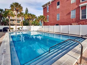 A swimming pool with a metal ladder, surrounded by a white fence, palm trees, and a red building in the background. - Gulf Front Corner Unit & Million Dollar Views~ Chb305 (Madeira Beach)