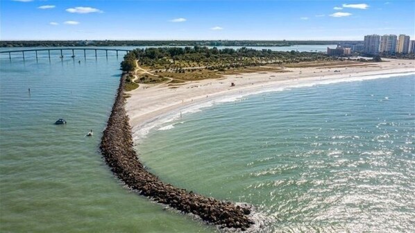 Enjoy a serene beach experience with a gentle curve leading to a rocky breakwater, surrounded by lush trees and the distant urban skyline.