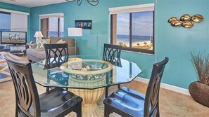 A dining area with a glass table and chairs, a sofa, a TV, and a view of the beach.