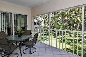 A balcony with a glass table and chairs, a potted plant, and a view of a green garden with trees.
