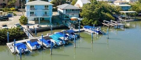 A marina with several boats docked, a building with a balcony, and a parking area with cars.