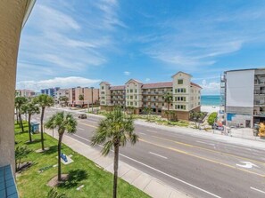 A beachfront hotel with multiple-story buildings, a road with palm trees, and a clear view of the ocean.