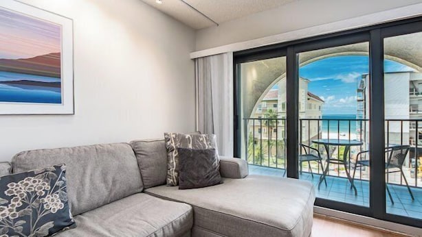 Ein modernes Wohnzimmer mit einem grauen Ecksofa, einem gerahmten Landschaftsbild und einer großen Schiebeglastür, die auf einen Balkon mit Gartenmöbeln führt.