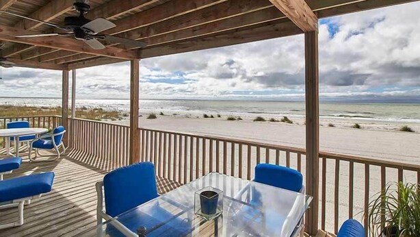 A covered outdoor seating area with a table and chairs, overlooking a beach with a cloudy sky.