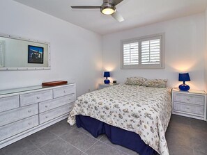 Une chambre à coucher comprenant un lit, deux tables de chevet avec des lampes, une commode, un ventilateur de plafond, une fenêtre avec des stores et un tableau encadré accroché au mur.