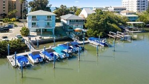A marina with several boats docked, a building with a balcony, and a parking area with cars.