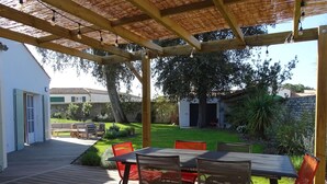 Een terras buiten, met een houten terras, een tafel met stoelen en een pergola met lichtslingers.
