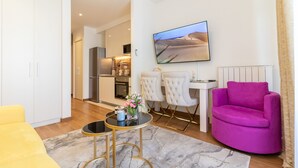 Una sala moderna con un sofá amarillo, un sillón rosa, una zona de comedor con sillas blancas y una cocina al fondo.