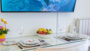 Una mesa de comedor preparada con platos, cubiertos, un bol de fruta y un vaso. Un televisor está montado sobre la mesa.