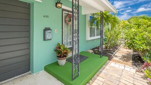 A house with a green exterior, a garage door, a front door with a wreath, a potted plant, and a pathway leading to the house. - Tropical oasis (Lake Worth)