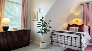 Ein Schlafzimmer mit einem Bett, einem Nachttisch mit Lampe, einer Topfpflanze und einem Fenster mit Vorhängen.