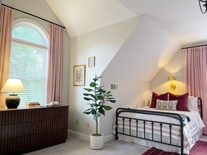 Een slaapkamer met een bed, een nachtkastje met een lamp, een potplant en een raam met gordijnen.