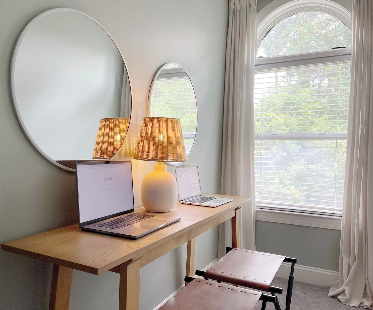 Une pièce avec un bureau, deux ordinateurs portables, une lampe et une fenêtre avec des rideaux.