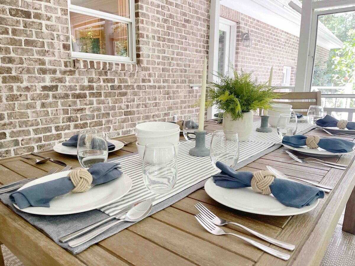 Une table à manger dressée avec des assiettes, des verres, des couverts et des serviettes, avec un mur de briques et une fenêtre en arrière-plan.