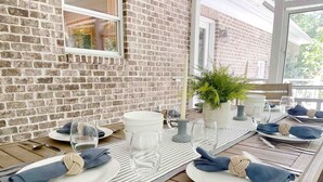 Ein Esstisch ist gedeckt mit Tellern, Gläsern, Besteck und Servietten, im Hintergrund eine Backsteinmauer und ein Fenster.
