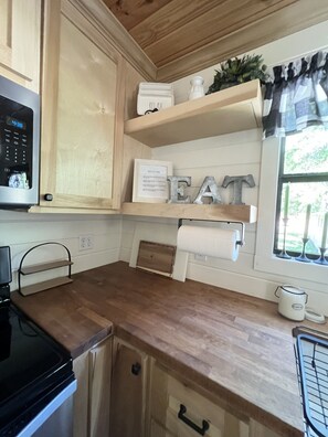 Una cocina con armarios de madera, microondas, cafetera y ventana con cortinas.