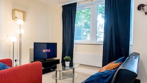 Un salón moderno con una televisión de pantalla plana, una mesa de centro de vidrio, sofás rojos y negros, y vistas al exterior a través de una ventana con cortinas oscuras.